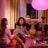 Two people sitting on a sofa with a colourful lamp above them, while another person sits at a table in the background.