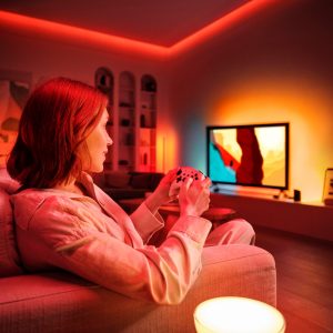 A woman plays video games and you can see immersive lighting on the wall behind the TV.