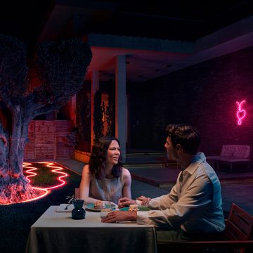 A couple dine on the patio with neon outdoor strip lights surrounding a tree and flowerbed and shining in pink tones of light. 