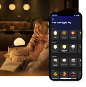 Person seated on a sofa holding a mug in a warmly lit living room, with a phone displaying Hue scene gallery options.