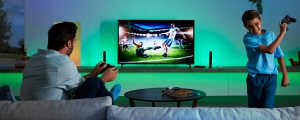 Two people playing video games in a living room with green smart lighting illuminating the TV and wall behind them.