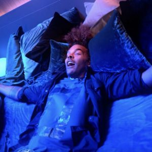 Person relaxing on a bed surrounded by pillows, with vibrant smart lighting casting blue and purple hues in a bedroom setting.