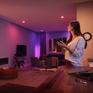 Person adjusting smart lighting with a mobile device in a modern living room, colored lights illuminating the space.