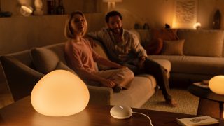 Two people sitting on a sofa in a living room, with a smart lamp and smart speaker emitting warm light on a table.