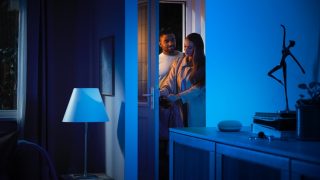 Two people enter a home illuminated by smart blue lighting, with a table lamp and smart speaker visible in the living room setting.