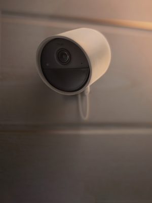 Wall-mounted smart security camera with cylindrical design, positioned on wooden panel, cable extending downward, lens facing outward.