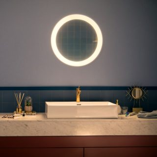 Round illuminated mirror mounted above a bathroom sink, casting light onto the marble countertop and gold faucet below.