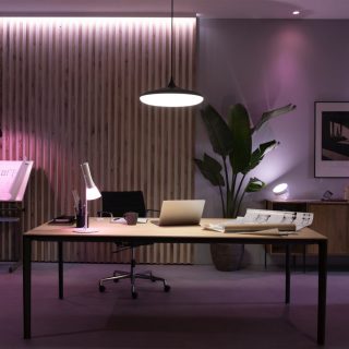 Pendant light fixture above office desk, casting bright illumination on workspace with laptop, lamp, and architectural drawings.
