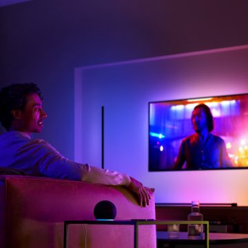 Homem em um sofá assistindo TV com iluminação ambiente e um alto-falante inteligente na mesa lateral