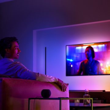 Homem num sofá a ver TV com iluminação envolvente e uma coluna inteligente na mesa de apoio